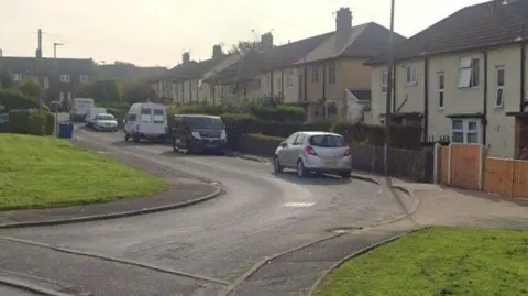Google Earth photograph of Pilmuir Road in Blackburn. Cars and vans are parked along the road, with a row of semi-detached houses and more homes at the top of the road. Two grass verges can be seen on either side of the entrance to the road.