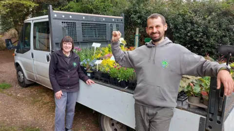 A man and a woman wearing branded hoodies stand next to a van which has a built-in trailer attached. The trailer is filled with potted flowers