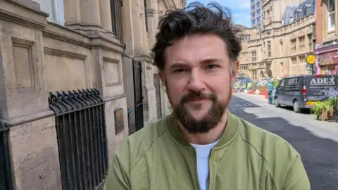 James Lovett, a man with a dark beard and hair is wearing a green jacket. He is standing outside Bristol Crown Court.