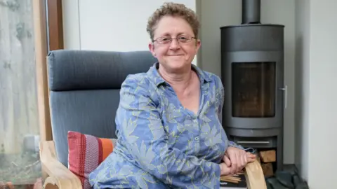 A woman sits in a grey armchair in a house. The woman has short curly hair and glasses, and she is wearing a blue shirt with yellow floral designs on it. There is a grey fireplace behind her on her right, and a large window behind her on her left.