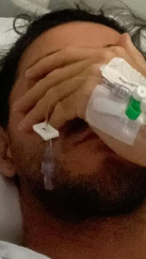 Man with a beard covers his face with is hand that has a medical tube attached.