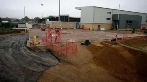 Northampton Town Football Club/ Peter Norton Behind the East Stand at Northampton Town, construction is on going. Big piles of dirt and the start of a tarmac road can be seen. The rest of the stadium is shown in the background. 