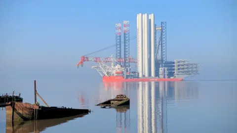 The Wind Peak sails along the Humber Estauary. The water is calm and the vessel is mirrored in the reflection of the water. The red and white vessel is carrying several large white towers and long wind turbine blades. Closer to shore, the submerged wreck of a small ship is breaking through the water.