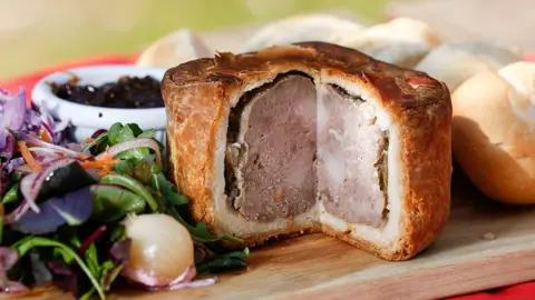 Getty Images/Bloomberg A photograph of a Melton Mowbray pork pie, with a slice missing, surrounded by salad to the left and a piece of bread to the right. 