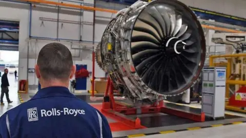 A Rolls-Royce worker looks on at a huge jet engine inside a factory