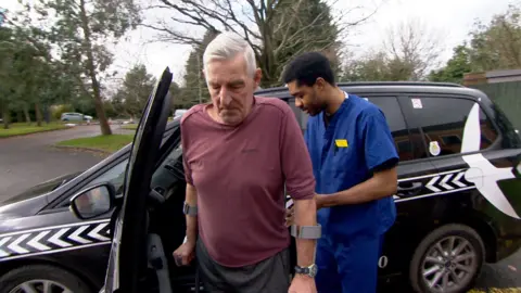 Nikki Fox/BBC A grey-haired man using crutches being helped into a waiting taxi by a man wearing surgical clothes