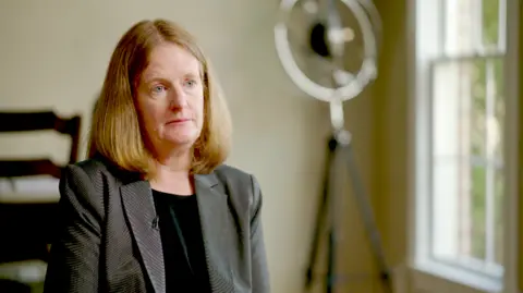 A middle-aged woman with straight auburn hair to her shoulder. She is wearing a formal grey jacket and black top. She is sitting in a room with a lamp, window and table in the background.