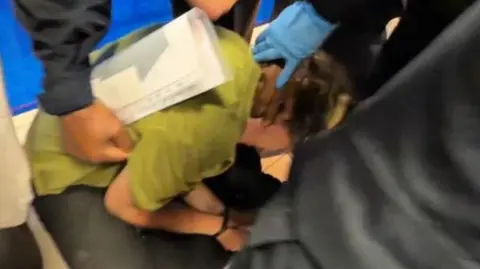 A woman in a green t-shirt is being forcefully pushed to the ground. She is on her knees with her hands cuffed. Three men have their hand on her back and head. 