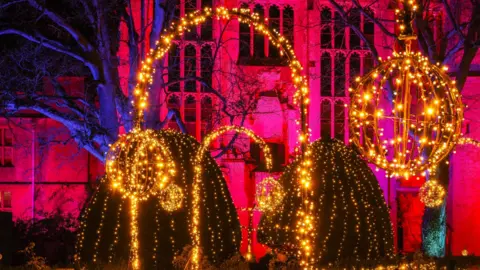 Steve Green A light spectacle, with an arch, two large baubles and two bushes, drapped in orange lights and the wall of a stately home lit up with pink light in the background