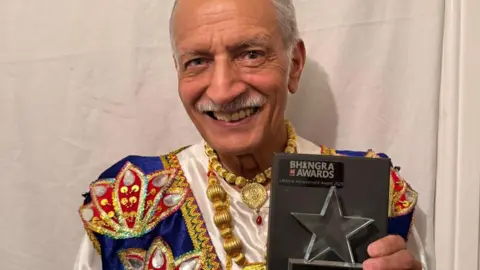 A man with short white-grey hair stands against a white backdrop. He is wearing a colourful white, gold, blue and red outfit and holding a grey award with a glass star