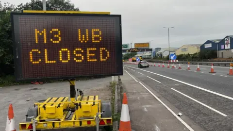 BBC A dual carriageway has orange and white cones running down the middle dividing line in the road. There is a large automated sign in the foreground that says 'M3 WB CLOSED'.