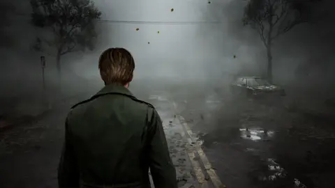 Konami A male character wearing an olive jacket is seen from behind as he walks into a very foggy, ominous scene. The view is obscured but road markings, two skeletal trees and an an abandoned car can be seen. Leaves, frozen in the air in this still, suggest a windy atmosphere. 