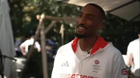 BBC Matt Hudson-Smith talking about his race. He is dressed in a white Team GB track suit and has a beard, moustache and gold earring.