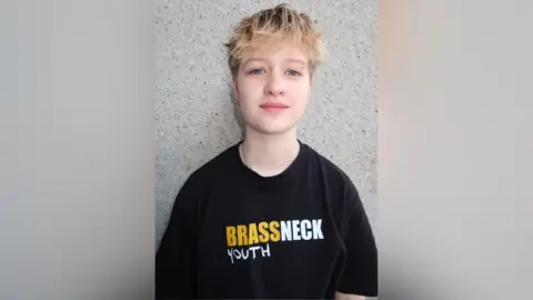 Brassneck Youth Theatre Fionn is wearing a black t-shirt with a yellow and white logo saying Brassneck Youth Theatre. He has a long blonde fringe and shirt hair. 

