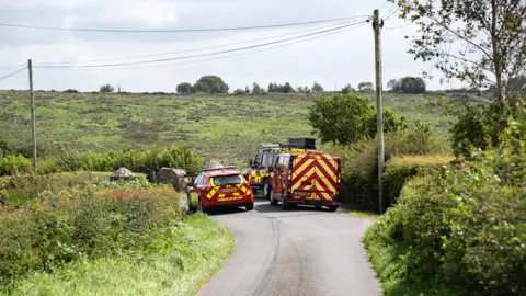 Pacemaker Red and yellow striped emergency vehicles are parked on a country lane which is surrounded by green fields.  