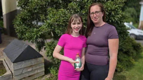 A girl in a pink dress next to her mum, with the girl holding a bottle of green washing up liquid