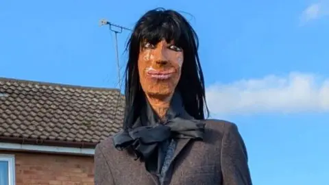 An effigy of Claudia Winkleman sitting on a garden fence.