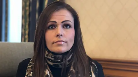 BBC Lucy Domaille - a woman with long brown hair, looking at the camera. She is wearing a leopard print scarf with a black jumper underneath. 