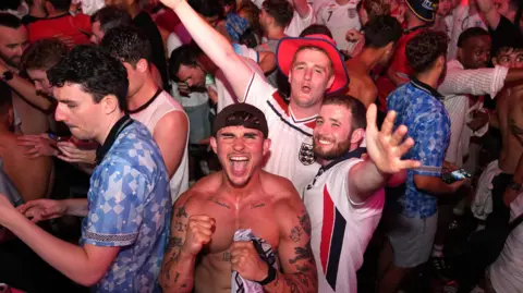 Three England supporters are smiling and celebrating while looking into the camera. This picture was taken in an event space, so there are lots of people celebrating with their friends. Must people are wearing the Blue and White England football kit.