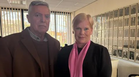Rachael O'Brien and her husband Joe. Mrs O'Brien is smiling into the camera and has short, blonde hair and is wearing a black top with a bright pink scarf around her neck. Her husband is standing to the left and has short, grey hair and is wearing a brown coat, shirt and a checked shirt. They are standing in a large room with has a wall covered in black and white portraits of people. There is a large window with vertical blinds behind them.