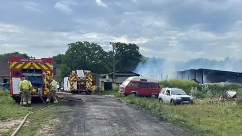 Qays Najm/BBC Fire crews are parked on a driveway which leads to outbuildings. The large building on the right has smoked coming from it and several cars parked in front of it.
