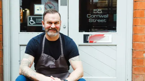 Claire Lishman PR Glynn Purnell in a black t-shirt and apron, sat outside a white door at the front of his restaurant in Birmingham.