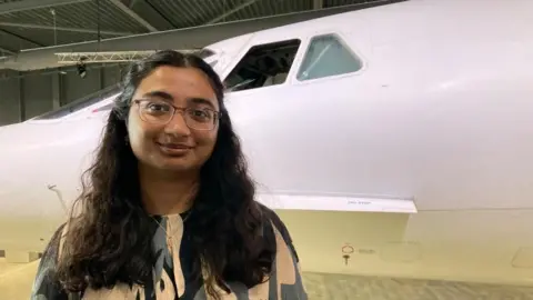 A woman with dark shoulder length hair smiles in front of the cockpit window of a concord jet on display at Aerospace Bristol