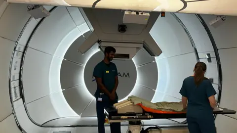 Sharon Barbour/BBC Inside the proton beam therapy room, two of the clinical team prepare the bed. The machine is large and lit up. It is circular and there are circles of light creating a tunnel effect. 