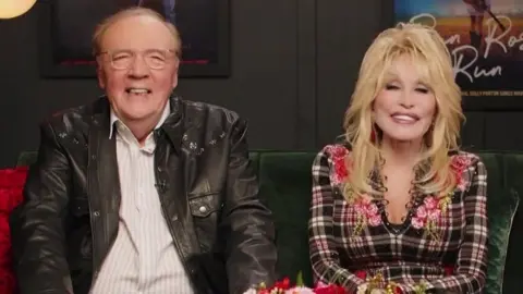 RTE James Patterson, seated to the left, has thin, receded greyish hair, and wears glasses, a shirt and leather jacket, and is smiling. Dolly Patron has blonde curled hair and is wearing a low-cut checkered dress and is smiling. 