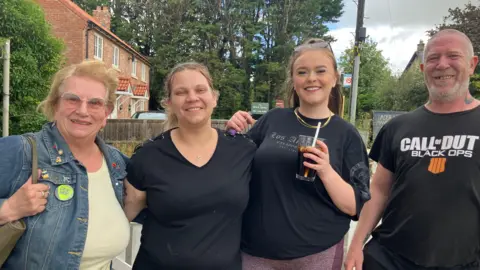 A woman in a denim jacket stands next to two women in black t-shirts and a man in a black T-shirt.