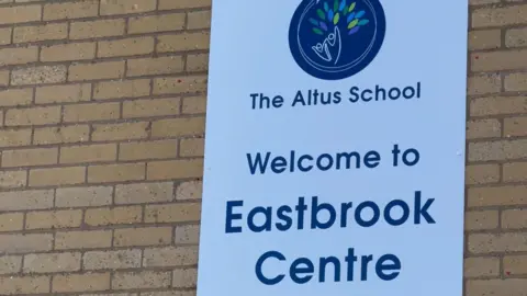 A brick wall with a sign which reads 'Altus School - Welcome to Eastbrook Centre'. The writing is in blue on a white sign. There is a logo depicting two cartoon figures holding hands and a number of leaves coloured in blue, light green, dark green and purple.