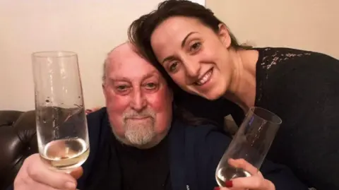 Natalie Cassidy Natalie leans in to her dad, who's sitting on a sofa, as they pose for the camera, both holding champagne flutes.