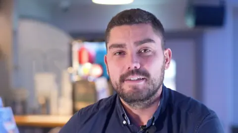 A close-up of a man with short dark hair and facial hair looks into the camera while sitting at what appears to be a bar - although that part of the image is blurred. He is wearing a navy shirt. 