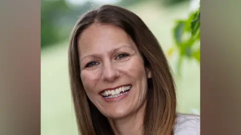 Kirstie Cook Kirstie Cook smiles at the camera while standing outside. She has long dark brown hair and is wearing a white top.