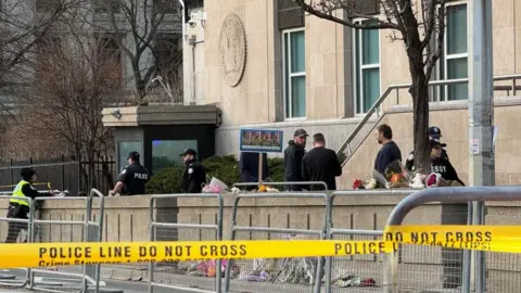 An image showing law enforcement members outside the US consulate building in Toronto, surrounded by caution tape. There is also a small memorial visible for the US soldiers killed in the Middle East during the ongoing war in Iran, with flowers laid around a sign with their photos.