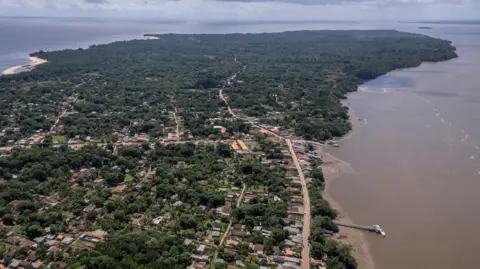 Anderson Coelho/Getty Images A spit of land jets out into the sea. The land is populated with trees, low lying house and roads winding through the forest.
