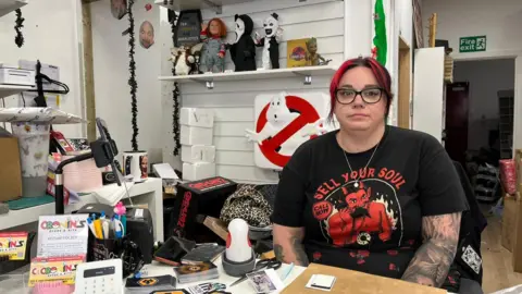 A woman with red hair and a black and red top sits behind a desk. 