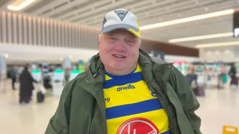 Mark smiles at the camera, he wears a green jacket over a blue and yellow striped Warrington shirt and a grey and yellow hat.