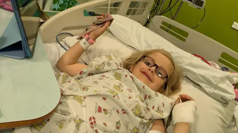 PA Media Sofia Hill lying in a hospital bed. She has blonde hair, glasses, a hospital gown on and a bandage on one hand. She is looking at the camera and smiling.