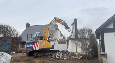 Jamie Niblock/BBC A large yellow digger tears down the roof of a home on a cloudy day. Rubble is on the ground around the house and digger.