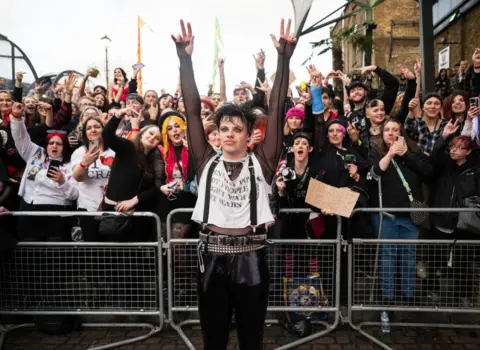 Samir Hussein/WireImage Yungblud stands in a white T-shirt with his hands in the air in front of a crowd of fans behind metal railings.