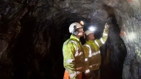 Dalradian Resources A cave with two men in orange high viz at work pointing at the wall  