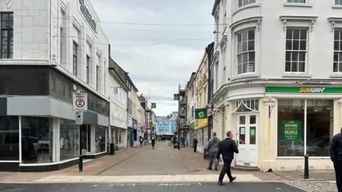 A view looking down King Street in Whitehaven. There is a Subway shop on the right with an empty unit on the left. Several signs look tired and dirty, with scaffolding visible further down the pedestrian street.