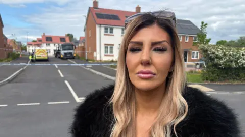 Kayleigh is stood near the police cordon and is wearing make-up, a black fluffy jacket or to. She has long brown/blonde hair and has her sunglasses on her head. Police tape and vehicles can be seen in the street behind her.
