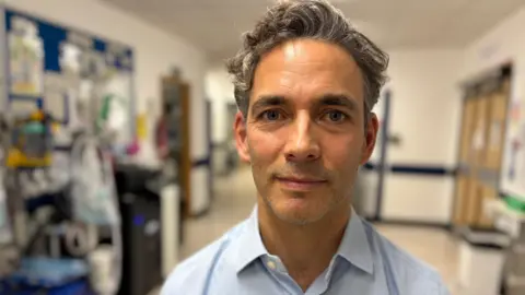 A man with short hair is wearing a light blue shirt and standing in a hospital corridor.