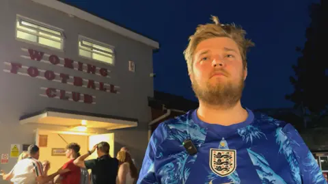 BBC / Zac Sherratt Ell Guidera wearing an England shirt stands outside the Woking Football Club building at night, with several people gathered near the entrance in the background.