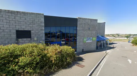 A Google street view image of the leisure centre in Saltash. A hedge is pictured in front of the building with a road running next to it. The building is made of grey brick and has large windows. There is a clear blue sky behind it as well as some white houses and green fields.