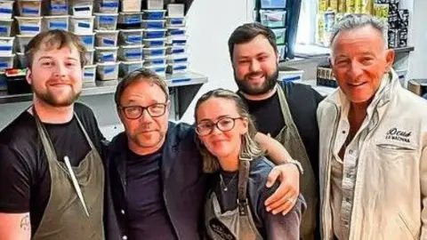 A group of people pose for a picture with actor Stephen Graham and Bruce Springsteen. Most of them are wearing brown aprons as they stand in a kitchen area.