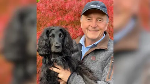 Colin J Butcher The image shows a man posing with his black dog; he is wearing a grey cap with "Musto" written on it, and a thick grey jacket. Behind them red leaves can be seen.