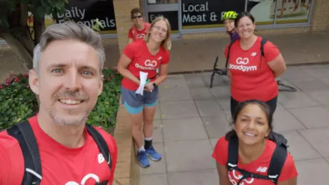 Ed Woollard A man and five women in red T shirts that read 'GoodGym' in white wearing fitness clothing standing on a street.